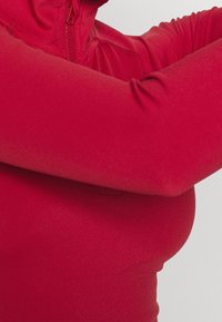 Rotes, eng anliegendes Sportoberteil mit langen Ärmeln, hohem Kragen und Viertel-Reißverschluss. Glatte Textur ohne sichtbare Muster oder Verzierungen.