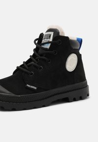 Botas de tornozelo em camurça preta com sola de borracha, apresentando detalhe em logótipo circular, puxador azul em contraste e atacadores pretos. Padrão texturizado na sola.