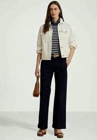 Kobieta w białej, krótkiej kurtce, w granatową górę w paski, szerokich czarnych spodniach, na koturnach, trzymająca brązową torebkę, stojąca na jednolitym tle.