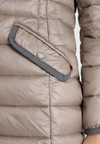 Béžová péřová bunda s diagonální kapsou. Měkký, lesklý materiál s šedými akcenty a stříbrným detailem knoflíku. Prošívaný design dodává texturu.
