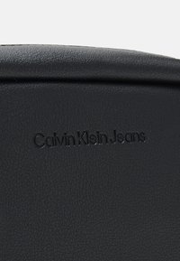 Superficie in pelle nera texture che presenta il testo in rilievo "Calvin Klein Jeans" in un font semplice. Design strutturato e minimalista.