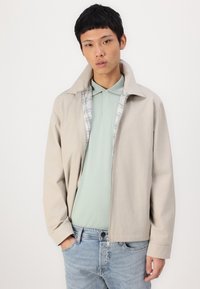 Veste légère beige avec un col, ouverte pour révéler un polo vert clair et une doublure à carreaux. Le modèle se tient avec une main dans la poche, portant un jean bleu.