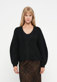 Cardigã preto de malha com decote em V e fecho de botão único, com mangas largas e um padrão texturizado, emparelhado com uma saia de estampa de leopardo.