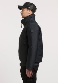 Mężczyzna w czarnej kurtce z wysokim kołnierzem i czapce baseballowej z siatki, stojący w profil na tle czystego białego tła.