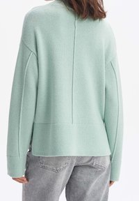 Mintunvihreä neulepusero, jossa on löysä istuvuus, korkea kaula-aukko, roikkuvat olkapäät ja ribattu hihansuut, yhdistettynä vaaleanharmaisiin farkkuihin.