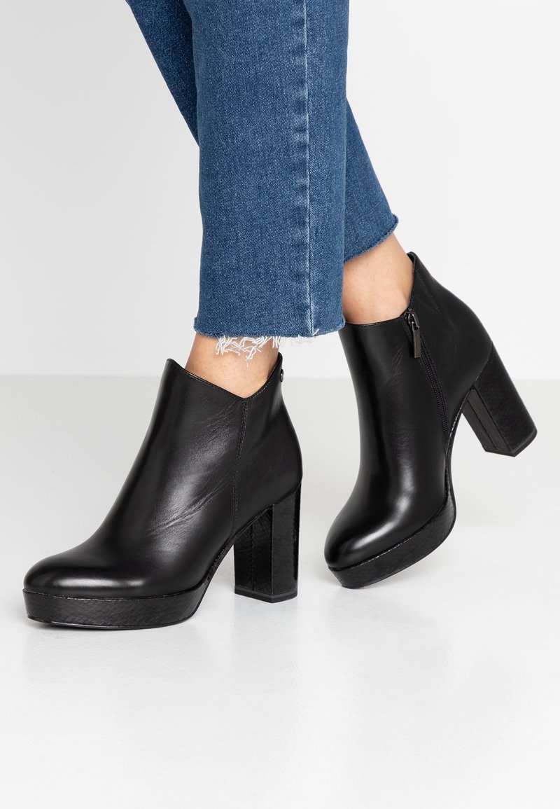 Bottines à plateforme en cuir noir avec fermeture éclair latérale, portées avec un jean bleu à ourlet effiloché, sur sol blanc et fond neutre.