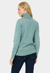 Gai aqua turtlenecks džemperis ar ribotu apkakli un aprocēm, gluda tekstūra un brīvs piegriezums, valkāts ar tumši ziliem džinsiem.