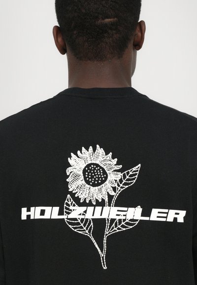 Tricou negru cu un design brodat alb al unei floarea-soarelui și textul "HOLZWIELER" dedesubt. Croială lejeră, cu decolteu rotund și mâneci scurte.