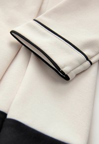 Tessuto beige con una finitura testurizzata, caratterizzato da un bordo nero sul polsino della manica. Il polsino è piegato, rivelando un design a strati.
