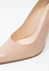 Chaussure à talon haut nude brillante avec bout pointu et intérieur en cuir lisse, présentée en gros plan sur fond blanc.