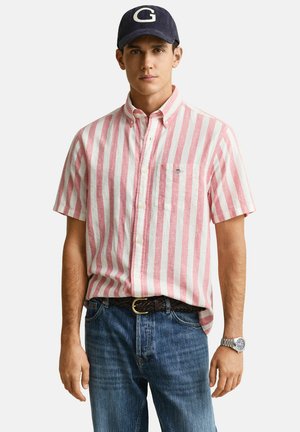 Homme portant une casquette bleu marine avec un "G" blanc, une chemise à manches courtes rayée verticalement rose et blanche, un jean bleu, une ceinture tressée et une montre-bracelet argentée.