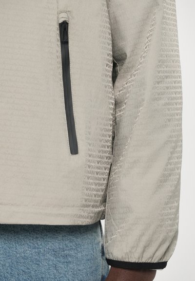 Helehall hall jakk, millel on õrn kogu ulatuses logomuster, lukuga küljetasku, elastsed mansetid ja sile tekstuur.