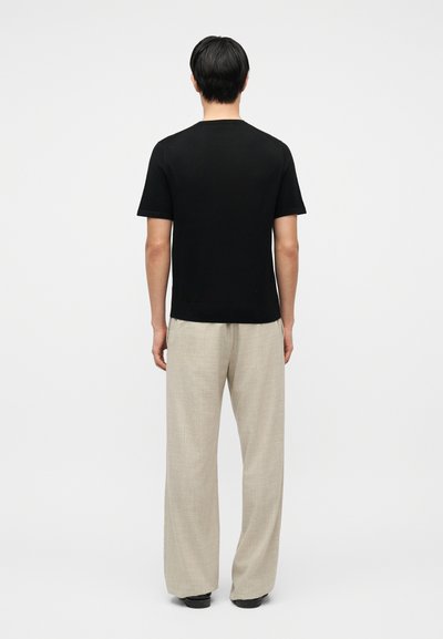 Άνδρας με μαύρα μαλλιά, που φοράει μαύρο κοντομάνικο μπλουζάκι και μπεζ παντελόνι με φαρδιές πιέτες, στέκεται γυρισμένος με την πλάτη προς το μέρος του θεατή, μπροστά σε ένα απλό φόντο.