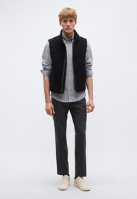 Fekete suede mellény világoskék gombos ing fölött, szürke szabott nadrág és bézs sportcipő, sima háttér előtt állva.