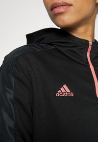 Sweatshirt à capuche noir avec des épaules en tissu noir mat et brillant contrastant. Il présente une fermeture éclair rose et un logo sur la poitrine.