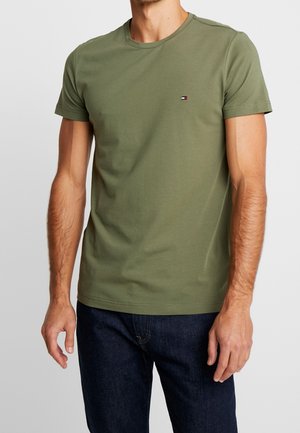 Mann trägt ein olivgrünes kurzärmliges T-Shirt mit Rundhalsausschnitt und kleinem rechteckigem Logo auf der linken Brust, kombiniert mit dunkelblauen Jeans.