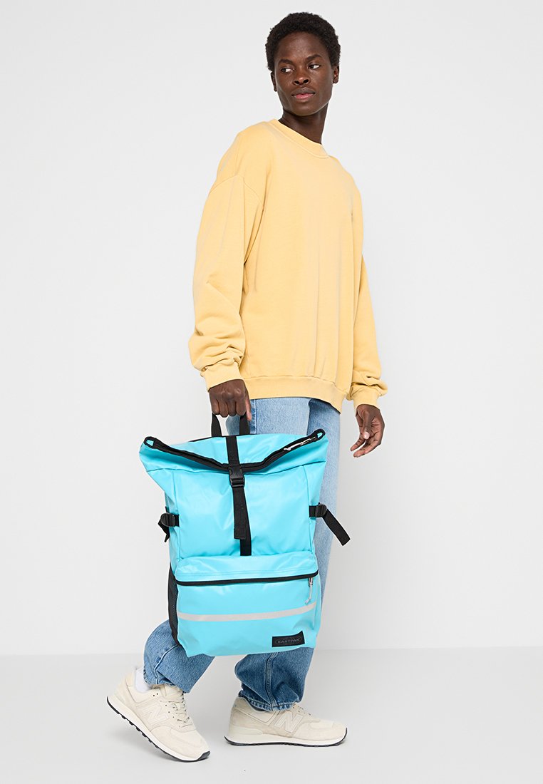 Hellblaue wasserfeste Rucksack mit schwarzen Trägern, faltbarem Deckel und einer Frontreißverschlusstasche. Das Modell trägt einen gelben Pullover und hellblaue Jeans.