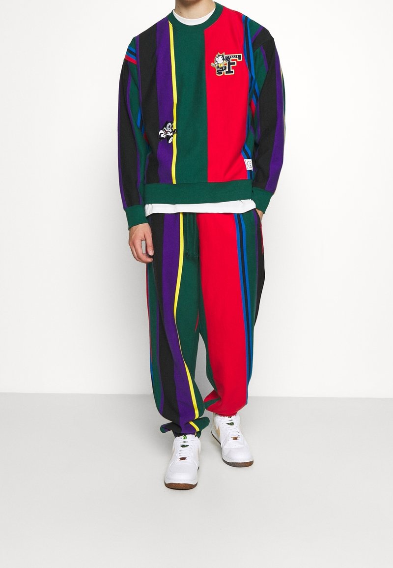 Personne portant un sweat-shirt rayé coloré et un pantalon assorti avec des patchs de dessins animés et des baskets blanches sur fond uni.