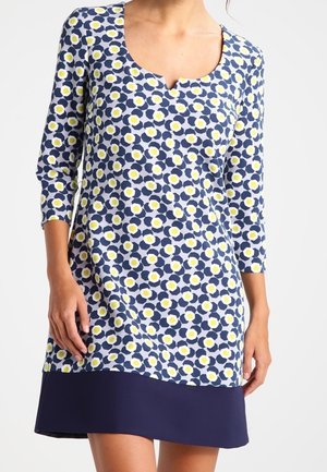 Femme portant une robe bleue avec un motif floral jaune et blanc, manches 3/4 et un ourlet bleu foncé uni.