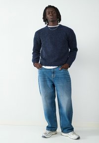 Granatowy sweter robiony na drutach z ściągaczami, luźne jasnoniebieskie dżinsy oraz szare sneakersy z siateczkowymi akcentami. Prosty detal w postaci srebrnego łańcuszka.