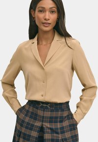 Camicia beige con bottoni e colletto, abbinata a una gonna a quadri blu navy con dettagli marroni e una cintura decorativa. Tessuto liscio.