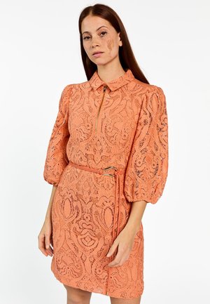 Frau mit langen Haaren, die ein korallenfarbenes Spitzenkleid mit Puffärmeln, einem Kragen, einem Frontreißverschluss und einem passenden Gürtel mit goldener Schnalle trägt.