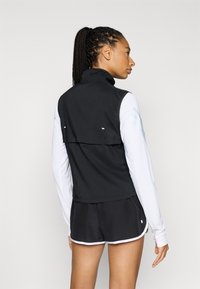 Svart ermeløs vest med høy krage, med klaff på ryggen og reflekterende detaljer, kombinert med hvit langermet topp og svarte shorts.