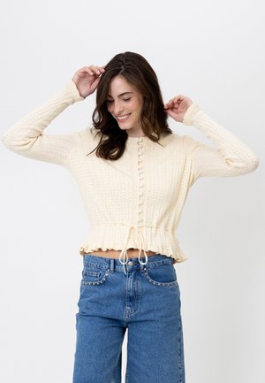 Giovane donna sorridente con le mani sollevate vicino alla testa, indossa un maglione a maglia color crema e jeans di denim blu su uno sfondo semplice.