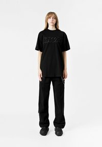 Camiseta oversized negra con un logotipo texturizado "MWM", combinada con pantalones sueltos negros con un acento de cadena y zuecos negros.