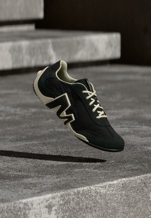 Sort og mørkegrønn joggesko med en blanding av glatte og strukturerte materialer, med hvit såle og karakteristisk sikksakkmønster på siden.