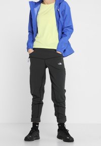 Blauwe waterdichte jas, lichtgroen shirt en zwarte tapered broek. Zwarte sneakers met gestructureerde zolen. Elastische manchetten op de jas.