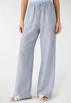 Donna con pantaloni a gamba larga a righe verticali azzurro chiaro e bianche, con vita con cordino, e sandali bianchi aperti sulle dita, in piedi contro uno sfondo bianco.