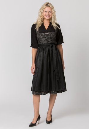 Blonde Frau lächelt, trägt ein schwarzes traditionelles Kleid mit gemustertem Oberteil, Puffärmeln, gebundener Schürze in der Taille und schwarzen High Heels.