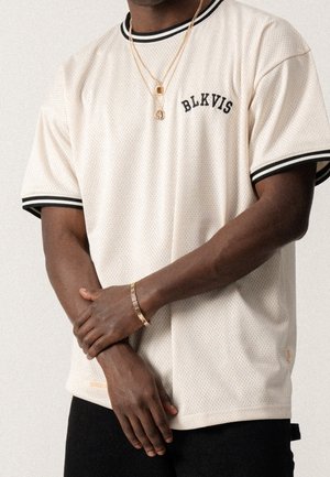 Homme portant une chemise en maille à manches courtes écrue avec des bordures noires et le texte "BLKVIS", des colliers en or et un bracelet en or, les bras croisés.