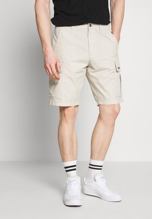 Beige cargo shorts with side pockets, made of cotton. Features a button closure and folded hem. Paired with white sneakers and striped socks.