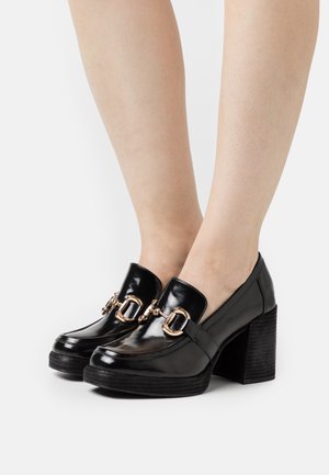 Zapatos de tacón de charol negro con un tacón grueso, que presentan acentos de hardware en tono dorado y un acabado liso y brillante.
