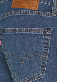 Jean en denim bleu avec une poche arrière présentant une couture incurvée distinctive, un patch en cuir beige avec les détails de la marque, et des surpiqûres dorées.