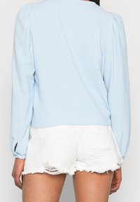 Blouse bleu clair à manches longues avec une finition texturée et des épaules bouffantes, associée à un short en jean blanc déchiré avec un ourlet effiloché.