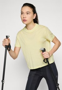Light yellow, short-sleeve shirt with a relaxed fit, paired with black leggings. The model holds hiking poles, showcasing a sporty design.