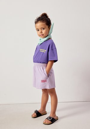 Enfant portant un t-shirt violet "American Vintage" et un short clair, debout les mains dans les poches, sandales noires et foulard vert clair autour du cou.