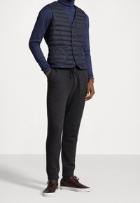 Marinblå quiltad väst med fyra knappar och två framfickor, tillsammans med mörkgrå tapered joggers och bruna snörskor.