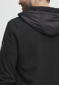 Crna dukserica s kontrastnom crnom, glatkom i blago sjajnom kapuljačom. Logotip na lijevoj rukavu. Mekana tekstura tkanine kroz cijelu duksericu.