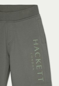 Jogging en coton gris avec une ceinture texturée, arborant un grand logo vert "HACKETT LONDON" sur le côté. Design lisse et décontracté.