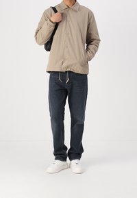 Beżowa lekka kurtka zapinana na guziki, z elastycznymi mankietami i ściągaczem u dołu, w połączeniu z ciemnoniebieskimi jeansami i białymi trampkami. Czarna torba na ramieniu.