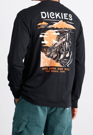 Personne portant un t-shirt noir à manches longues avec un motif de loup des falaises en orange et blanc et texte "Dickies Hard Work Since 1922 Fort Worth, Texas".