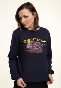 Felpa blu navy con una grafica frontale di una macchina d'epoca, testo "NEWPORT BEACH" e "ORANGE COUNTY SPEED SHOP". Vestibilità regolare con maniche lunghe.