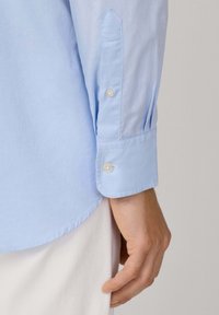 Camisa de manga larga de color azul claro con una tela texturizada. Los detalles notables incluyen un puño abotonado con dos botones y un dobladillo redondeado.