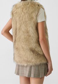 Brązowa kamizelka z futerka ekologicznego z wysokim kołnierzem, bez rękawów, o miękkiej fakturze, w zestawieniu z plisowaną spódnicą w kratę w kolorach beżowym i brązowym.