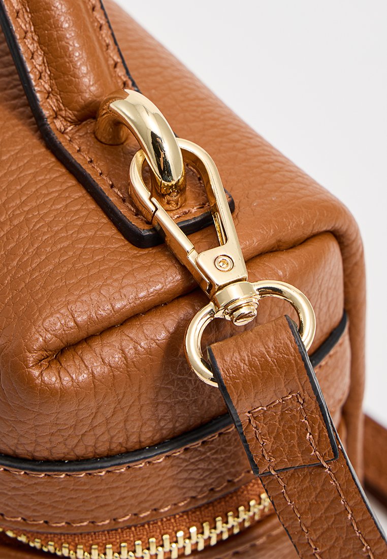 Détail d'un sac en cuir texturé marron avec une boucle en métal doré et une fermeture éclair, orné d'une liseré noir le long du bord de la sangle.