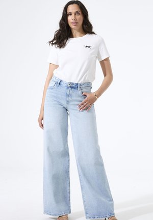 Frau trägt weißes T-Shirt mit kleinem Tiger-Design und hellblaue Jeans mit weitem Bein, steht vor einem schlichten weißen Hintergrund.